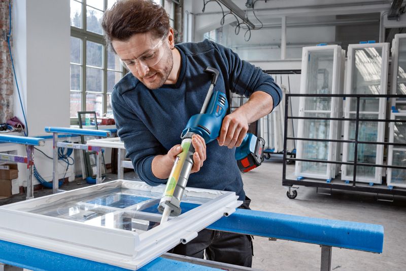 안전장비를 착용한 사람이 무선 코킹건을 사용하여 창틀에 실런트를 바르고 있습니다.