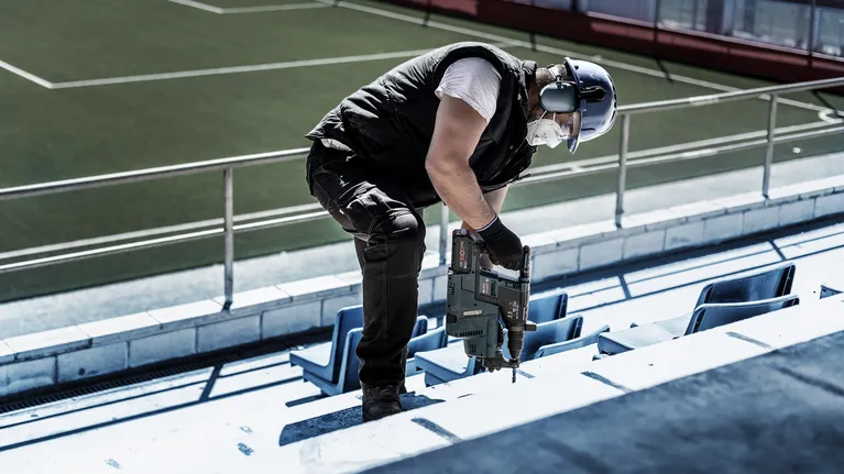 안전장비를 착용한 사람이 콘크리트 경기장 계단에 드릴을 박고 있습니다.