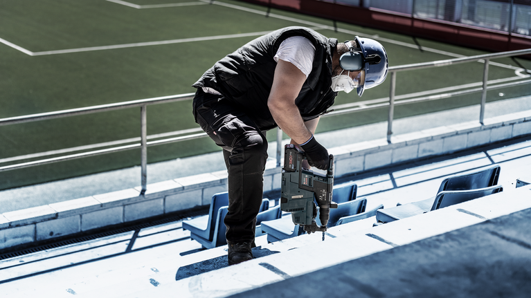 안전장비를 착용한 사람이 콘크리트 경기장 계단에 드릴을 박고 있습니다.