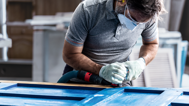 안전장비를 착용한 사람이 전동공구를 이용해 파란색 나무 표면을 연마하고 있습니다.