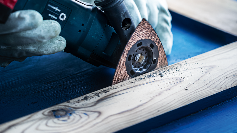 안전 장비를 착용한 사람이 멀티툴을 이용해 나무 판자를 연마하고 있습니다.