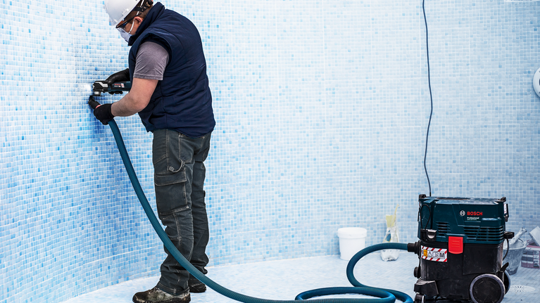 안전 장비를 착용한 사람이 진공청소기가 부착된 드릴로 수영장 타일에 구멍을 뚫고 있습니다.
