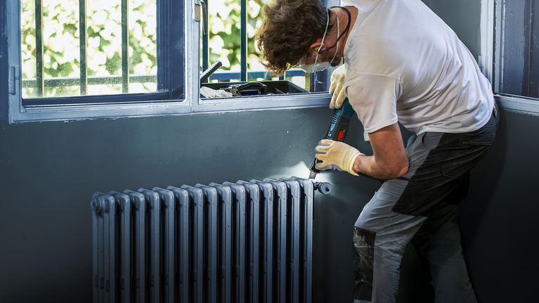 안전 장비를 착용한 사람이 무선 드릴을 사용하여 라디에이터를 설치합니다.