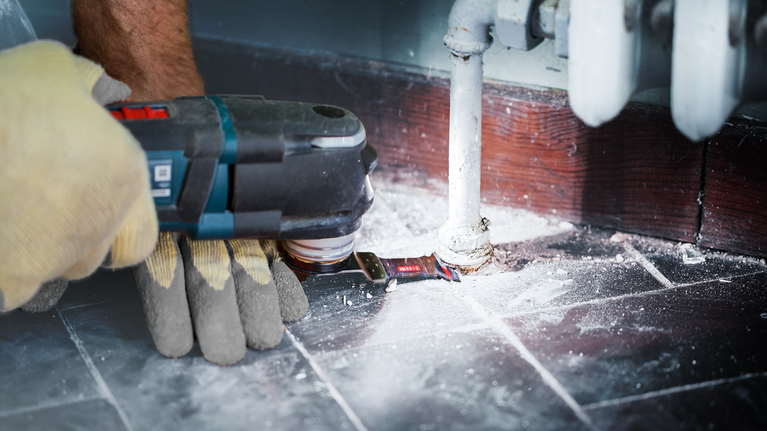 EXPERT MetalMax AIZ 45 AIT 멀티툴 블레이드 45mm 안전 장비를 착용한 사람이 전동 공구를 사용하여 파이프 주변의 바닥을 자르고 있습니다.