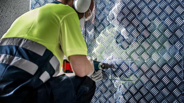 EXPERT MultiMax PAII 52 APIT 멀티툴 블레이드 52mm 안전 장비를 착용한 사람이 금속 무늬 벽 패널에 드릴을 박고 있습니다.