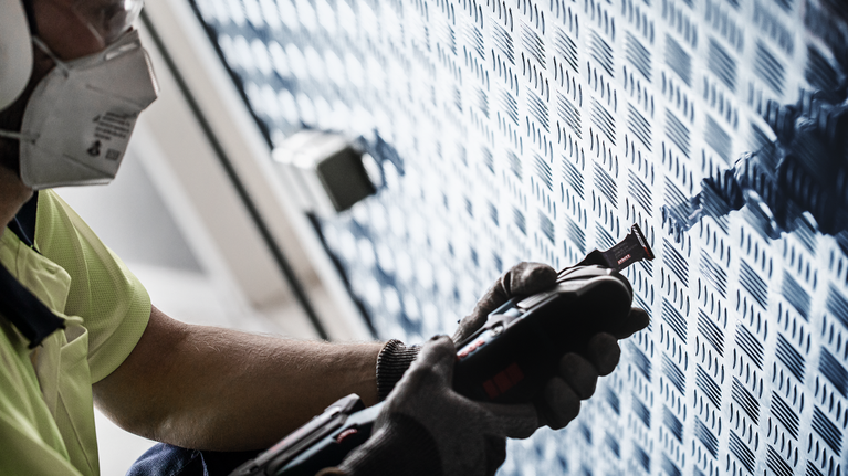 EXPERT MultiMax PAII 52 APIT 멀티툴 블레이드 52mm 안전 장비를 착용한 사람이 전동 공구를 사용해 무늬가 있는 금속 벽을 자릅니다.