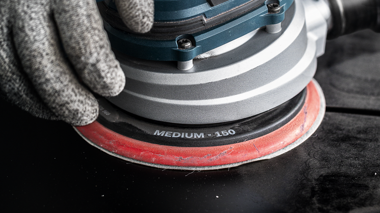 EXPERT Multihole 샌딩 패드, 범용, 150mm, 소프트 안전 장비를 착용한 사람이 어두운 표면에서 중간 입자의 오비탈 샌더를 조작하고 있습니다.
