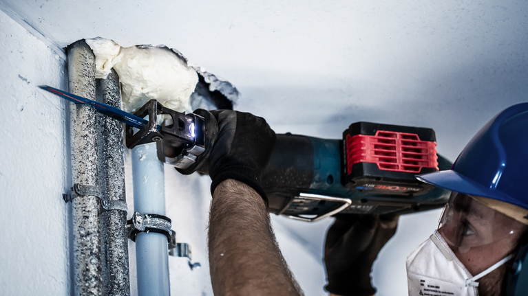 안전 장비를 착용한 사람이 왕복톱을 사용하여 금속 파이프를 자릅니다.