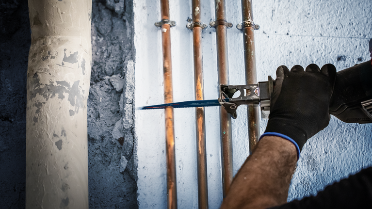 벽에 설치된 구리 파이프를 자르기 위해 왕복톱을 사용하는 사람.