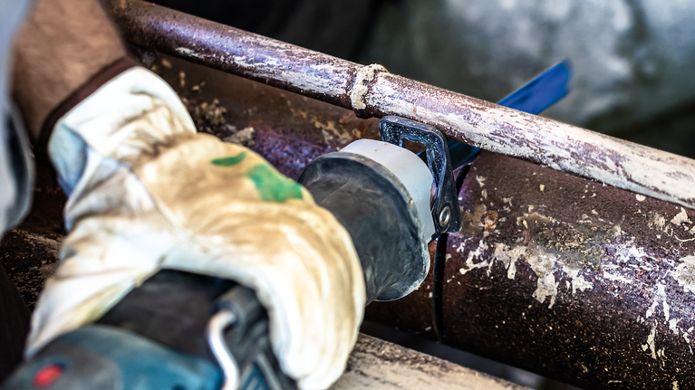 안전 장비를 착용한 사람이 왕복톱으로 녹슨 금속 파이프를 자릅니다.