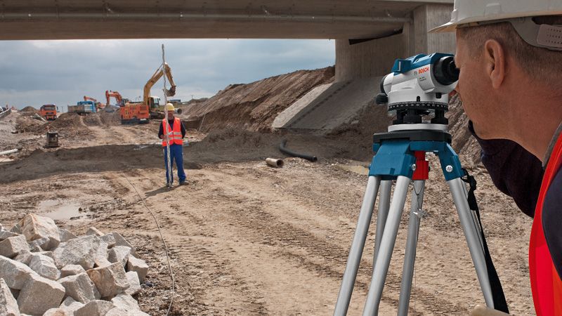 안전 장비를 착용한 사람이 도로 공사 현장에서 광학 수준을 사용하고 있습니다.