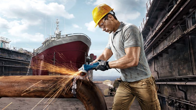 안전 장비를 착용한 사람이 조선소에서 금속 파이프를 갈아서 불꽃이 튀는 모습.