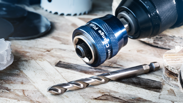 나무 표면 위에 금속 드릴 비트가 달린 드릴 척 부착물의 클로즈업입니다.