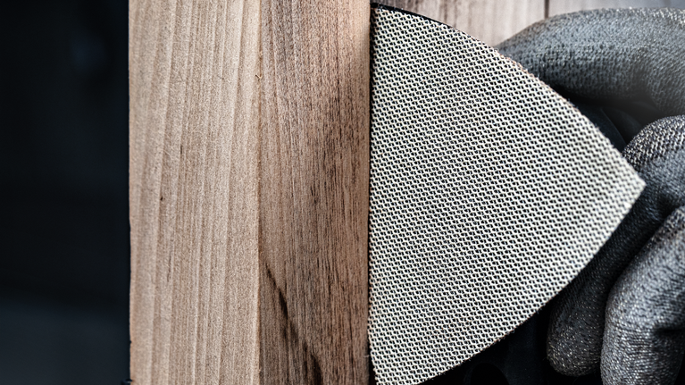 안전 장비를 착용한 사람이 메쉬 연마 패드를 이용해 나무 표면을 연마하고 있습니다.