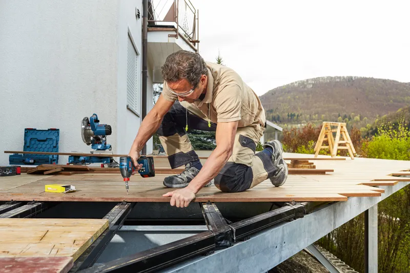 안전장비를 착용한 사람이 야외 파티오 프레임에 데킹 보드를 뚫고 있습니다.