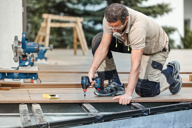 안전장비를 착용한 사람이 전동 드릴을 사용하여 야외에 데크 보드를 고정하고 있습니다.
