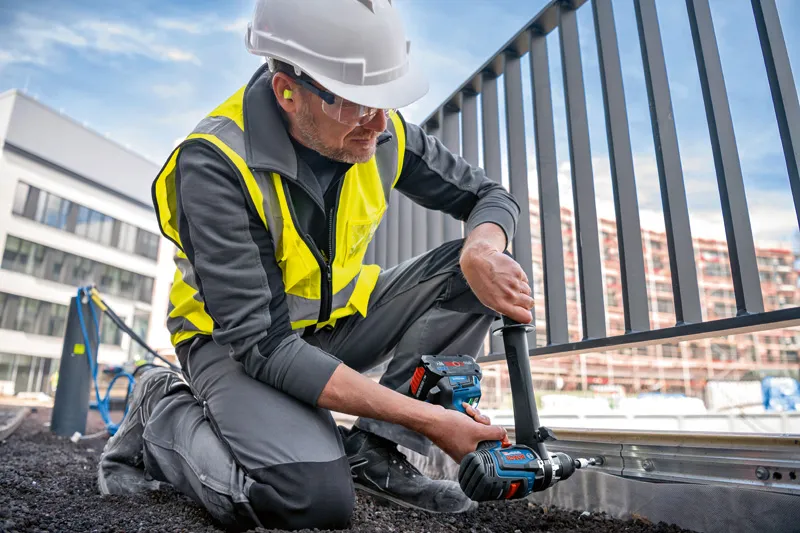 GSB 18V-150 C 안전 장비를 착용한 사람이 금속 울타리 프레임에 무선 콤비 드릴을 사용하고 있습니다.