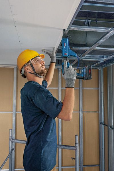 안전장비를 착용한 사람이 전동공구를 이용해 금속 천장 틀을 고정하고 있습니다.