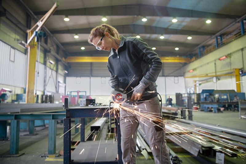 안전 장비를 착용한 사람이 공장에서 각도 분쇄기를 사용하여 금속을 절단하고 있습니다.