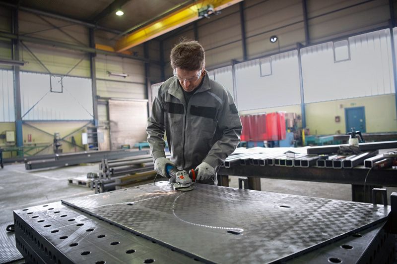 안전 장비를 착용한 사람이 공장 작업 공간에서 큰 금속판을 갈고 있습니다.