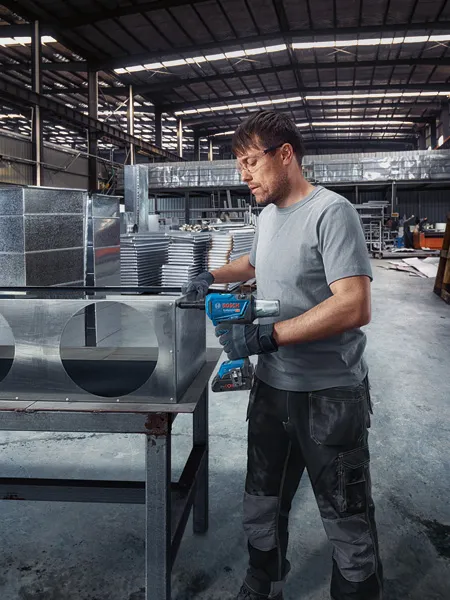 안전장비를 착용한 사람이 무선 리벳 건을 이용해 금속 배관을 고정하고 있습니다.