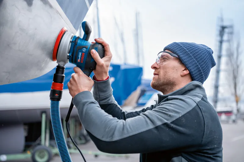 안전장비를 착용한 사람이 랜덤 오빗 샌더로 보트 선체를 샌딩하고 있습니다.