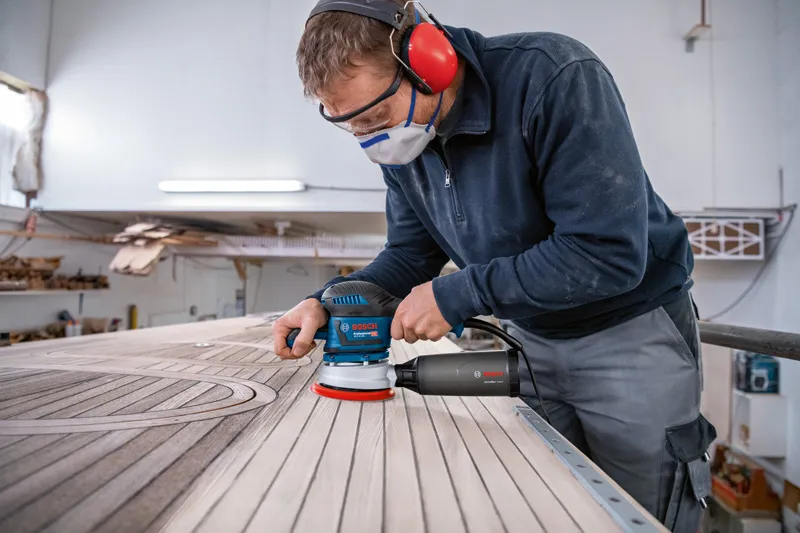 안전장비를 착용한 사람이 랜덤 오빗 샌더로 나무 판자를 연마하고 있습니다.