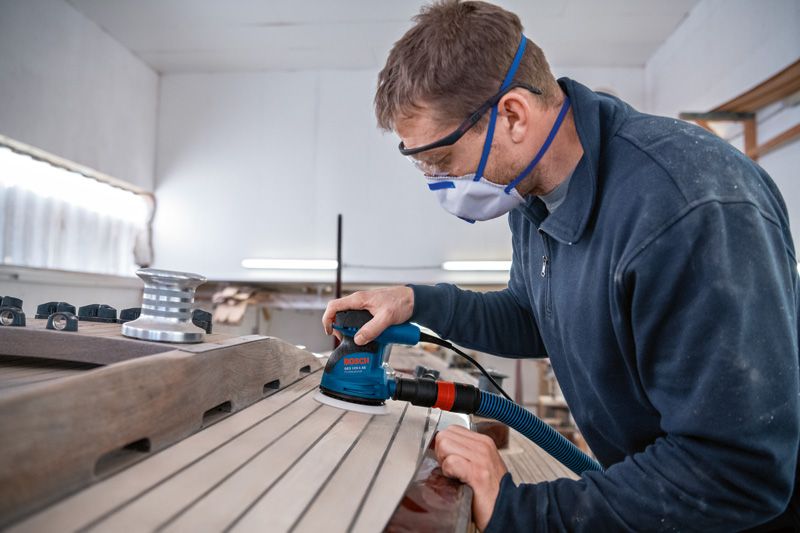 안전장비를 착용한 사람이 랜덤 오빗 샌더로 나무 판자를 연마하고 있습니다.