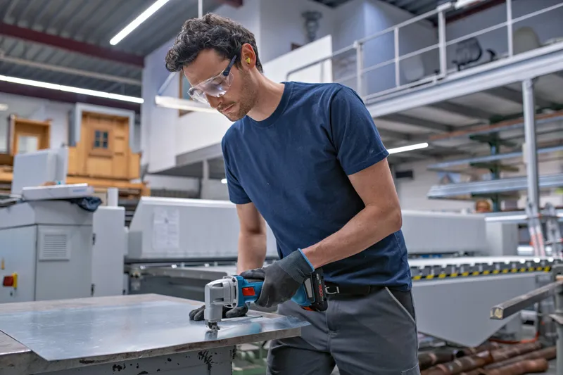 안전 장비를 착용한 사람이 무선 금속 가위로 금속판을 자르고 있습니다.