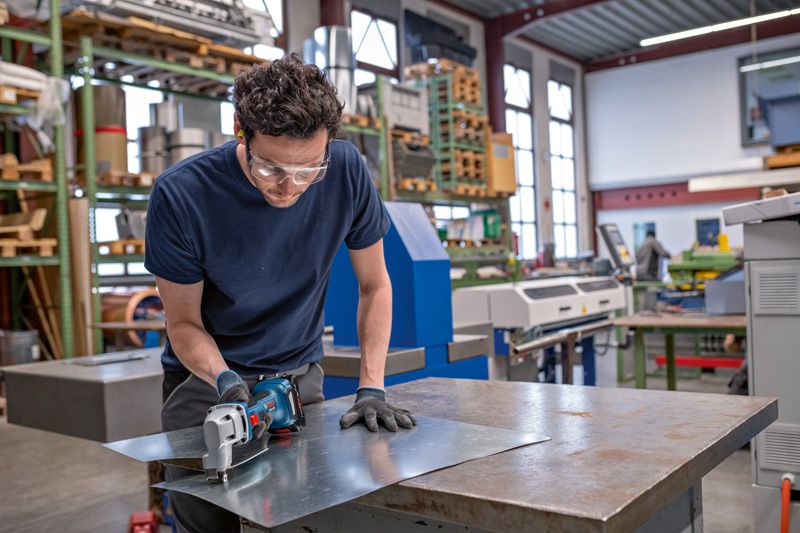 안전 장비를 착용한 사람이 무선 금속 가위로 금속판을 자르고 있습니다.