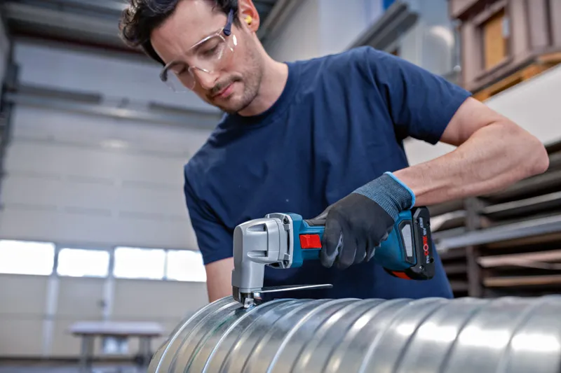 안전 장비를 착용한 사람이 무선 금속 가위로 금속 덕트를 절단하고 있습니다.