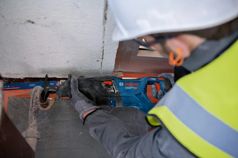 안전 장비를 착용한 사람이 왕복톱을 사용해 금속 파이프를 자릅니다.