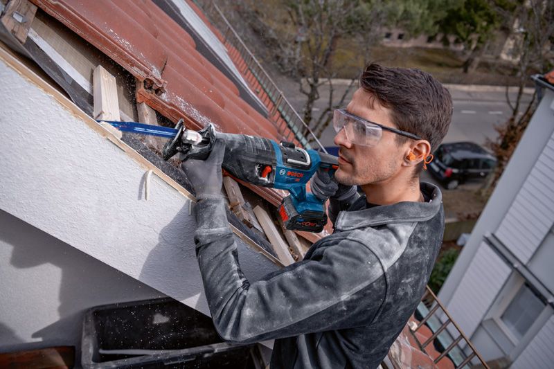 안전 장비를 착용한 사람이 왕복톱을 사용하여 야외에서 지붕용 목재를 자릅니다.