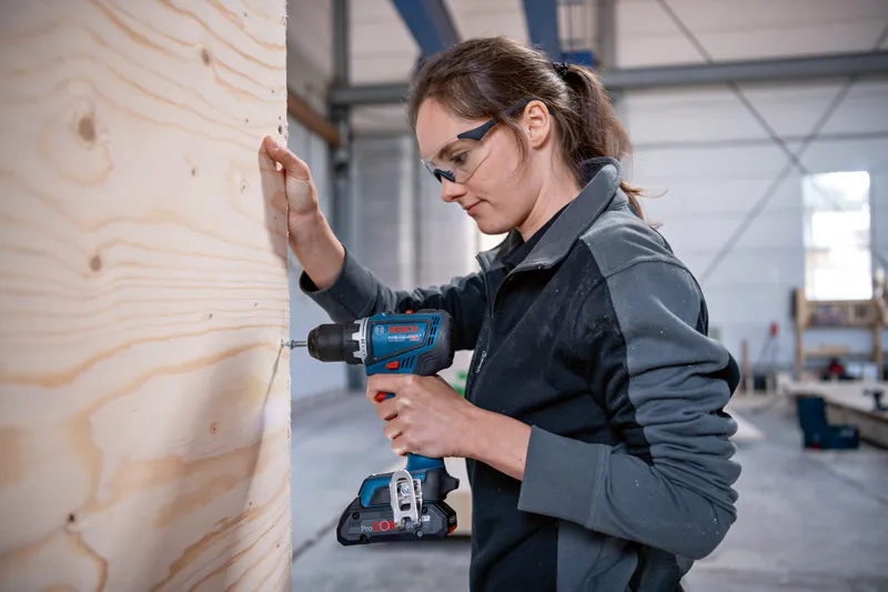 안전 장비를 착용한 사람이 작업장에서 나무판에 구멍을 뚫고 있다.
