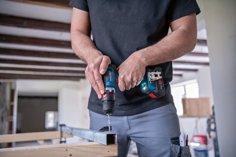 한 사람이 작업장 안에서 무선 드라이버를 이용해 금속 빔에 구멍을 뚫고 있다.