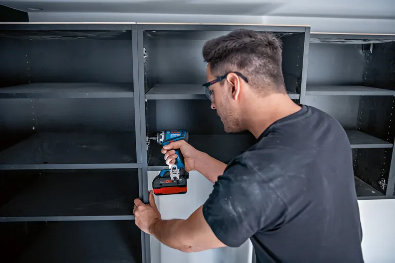 안전 장비를 착용한 사람이 무선 드릴이나 드라이버를 사용하여 선반을 조립합니다.