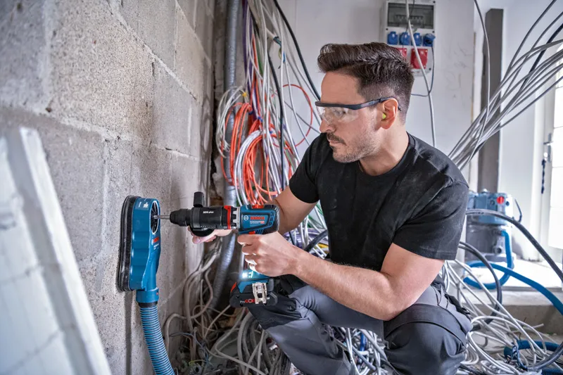 안전장비를 착용한 사람이 먼지 추출 장비를 이용해 콘크리트 벽에 구멍을 뚫고 있습니다.