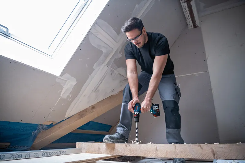 안전 장비를 착용한 사람이 반쯤 완성된 다락방에서 나무 들보에 구멍을 뚫고 있습니다.