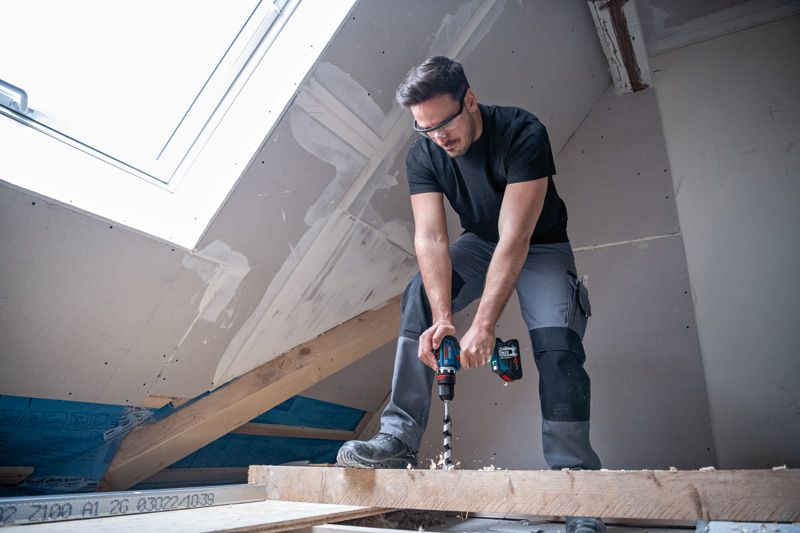 안전 장비를 착용한 사람이 반쯤 완성된 다락방에서 나무 들보에 구멍을 뚫고 있습니다.