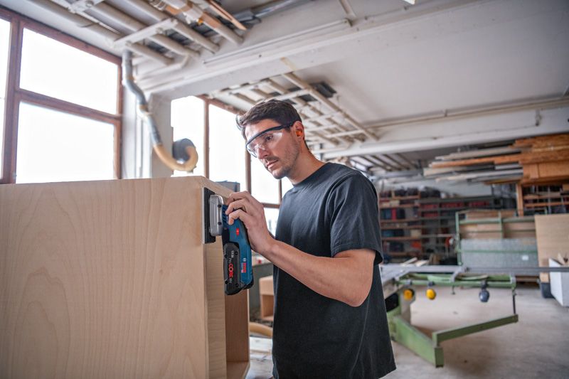 안전장비를 착용한 사람이 무선 오비탈 샌더로 나무 캐비닛을 샌딩하고 있습니다.