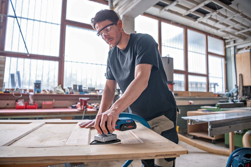 안전장비를 착용한 사람이 무선 오비탈 샌더로 나무 문을 샌딩하고 있습니다.