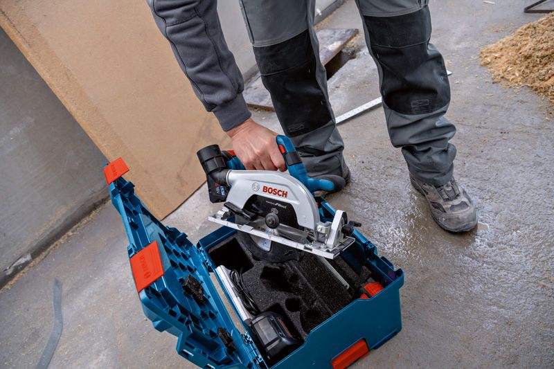 안전 장비를 착용한 사람이 작업장 바닥에 있는 도구 상자에서 원형 톱을 들어올리고 있습니다.