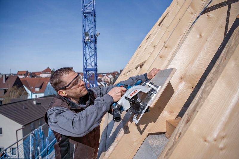 안전 장비를 착용한 사람이 원형 톱을 이용해 경사진 지붕의 나무판을 자릅니다.