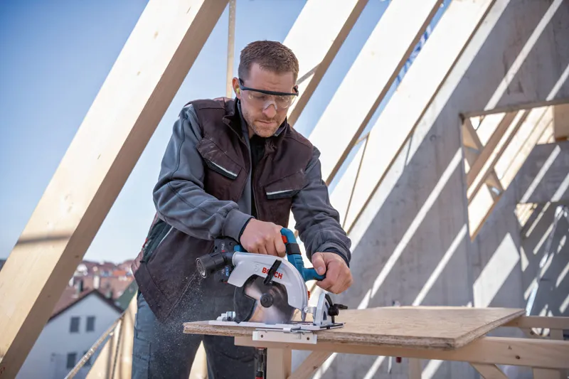 안전 장비를 착용한 사람이 건설 현장에서 원형 톱을 사용하여 합판을 자릅니다.