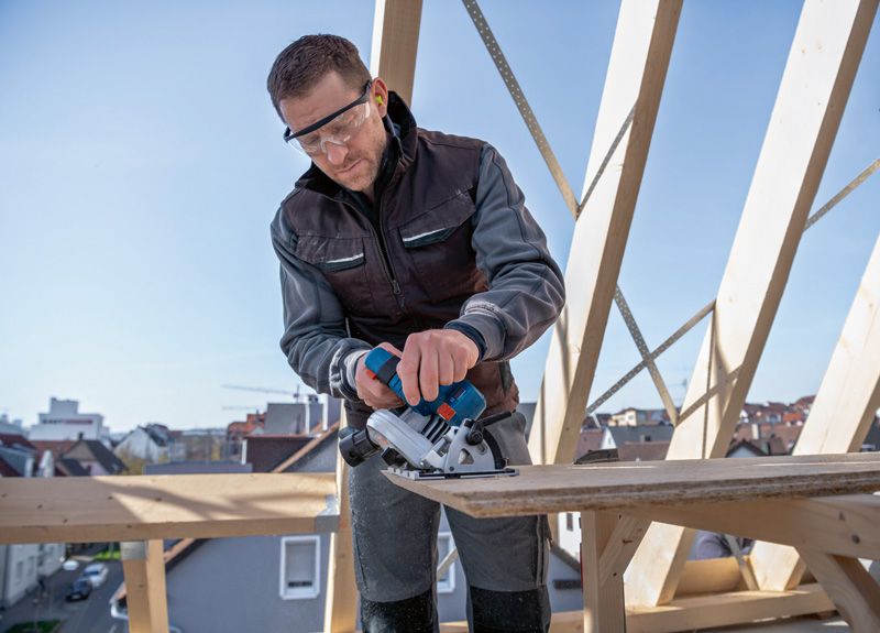 안전 장비를 착용한 사람이 건설 현장에서 무선 원형 톱을 이용해 나무를 자르고 있습니다.