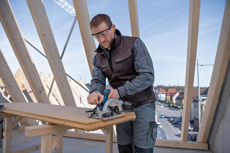 안전 장비를 착용한 사람이 건설 현장에서 원형 톱을 이용해 나무판을 자르고 있습니다.