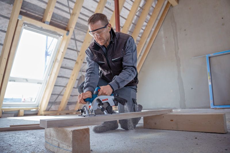 안전 장비를 착용한 사람이 미완성 다락방에서 원형 톱을 이용해 나무 판자를 자르고 있습니다.