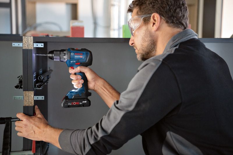 안전 장비를 착용한 사람이 무선 전동 드릴로 캐비닛 프레임에 구멍을 뚫고 있습니다.