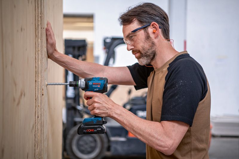 GDS 18V-330 HC 안전장비를 착용한 사람이 무선 드릴을 사용해 합판에 나사를 박고 있습니다.