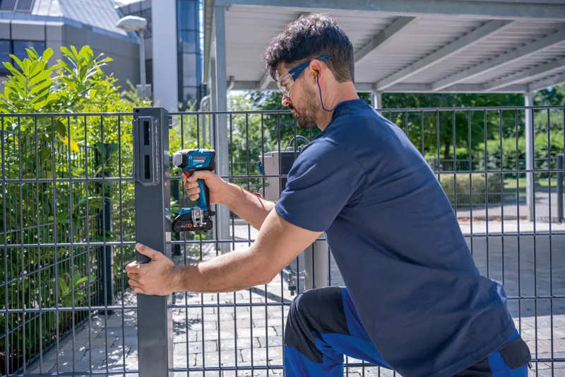 안전장비를 착용한 사람이 무선 드릴을 사용하여 금속 울타리 기둥을 고정하고 있습니다.